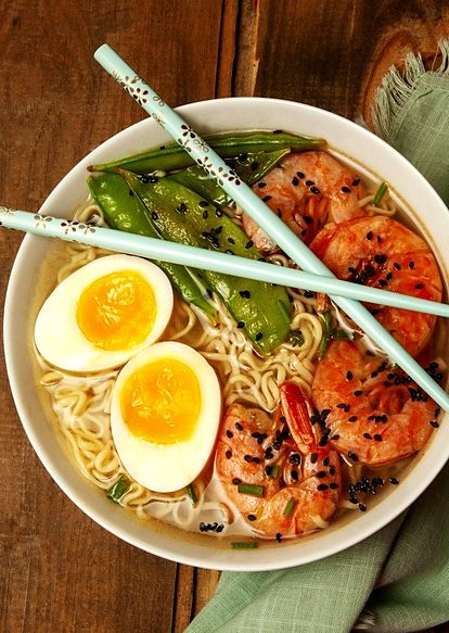 Bowl of ramen with shrimp, soft-boiled eggs, and snap peas, chopsticks resting across the rim